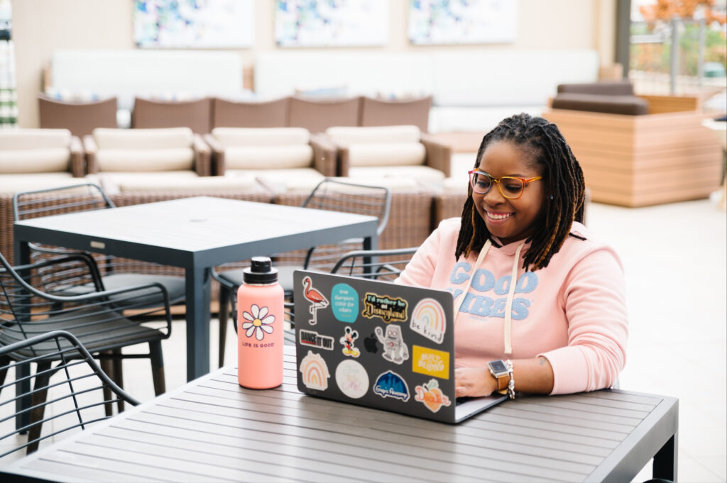 African American woman sitting outside at a table typing on laptop smiling.